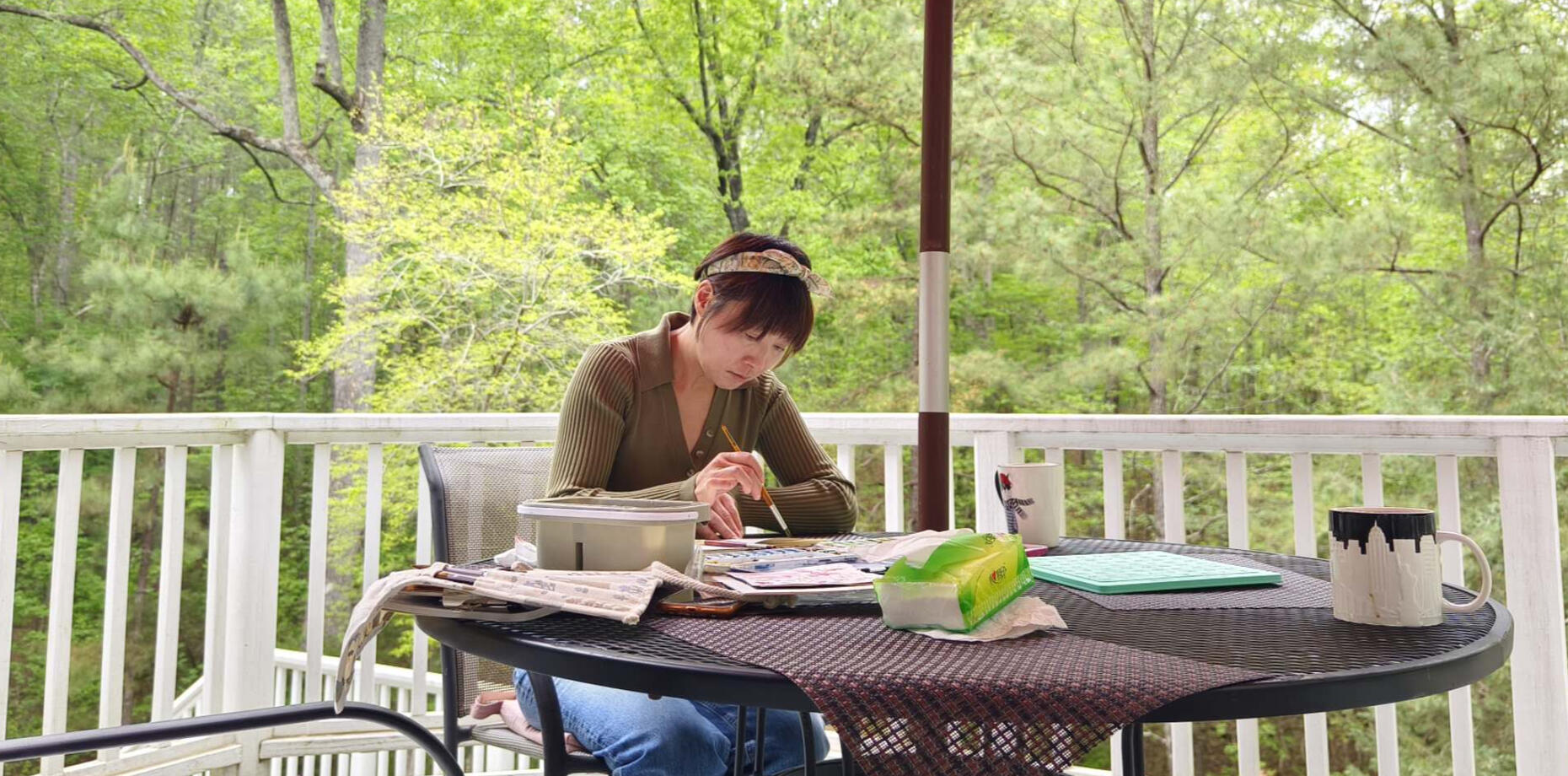 Artist Sherry Cheng painting watercolor outdoors on a porch, surrounded by spring greenery